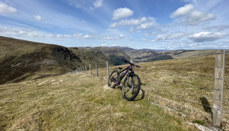A purple mountain bike resting against a wire fence on a grassy hillside, overlooking a broad valley under a blue sky with white clouds.