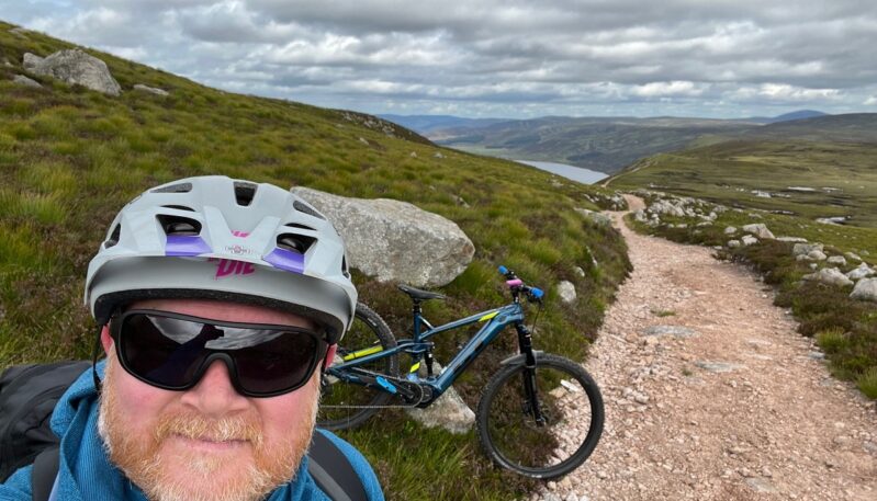 A selfie of a man in a helmet and sunglasses on a mountainside trail with a blue and yellow mountain bike behind him, overlooking a loch (Loch Muick) in the distance.
