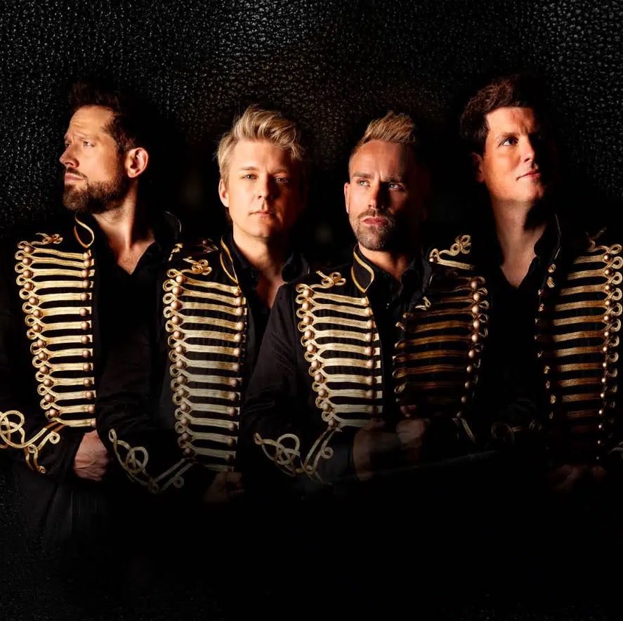 Four men in black shirts and black jackets with gold braid detailing, standing close together against a dark background.