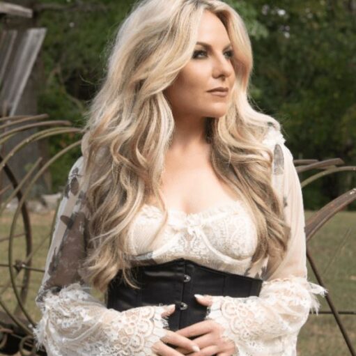 A blonde woman with long, wavy hair is shown outdoors, wearing a white lace dress with a corset-style black belt, looking off to the right.
