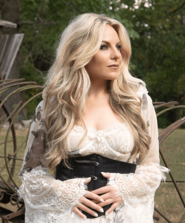 A blonde woman with long, wavy hair is shown outdoors, wearing a white lace dress with a corset-style black belt, looking off to the right.