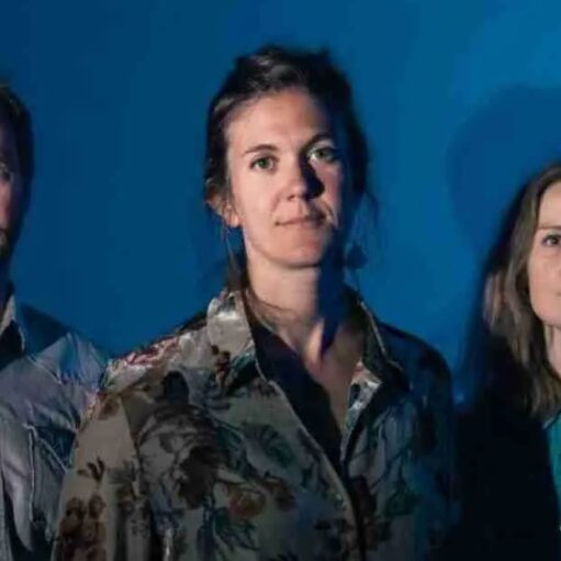 A man with a beard and two women, one with a floral shirt and the other in a blue shirt, stand close together against a solid blue background.