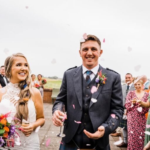 A smiling bride and groom are featured in the foreground after their wedding, with pink confetti petals falling around them, surrounded by applauding and smiling guests. The groom is wearing a grey suit jacket over a kilt, and the bride is wearing a high-neck lace dress and holding a bright bouquet.