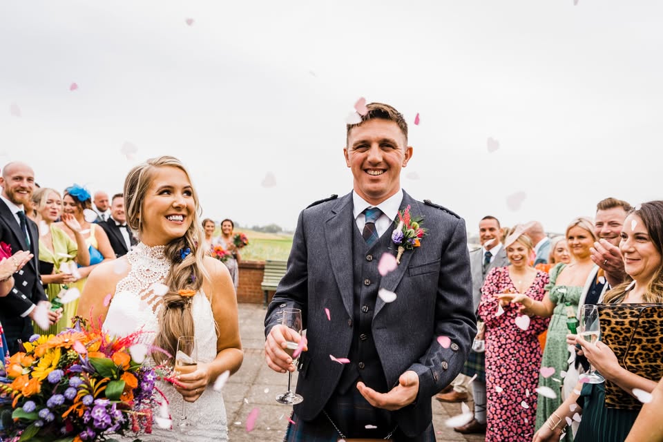 A smiling bride and groom are featured in the foreground after their wedding, with pink confetti petals falling around them, surrounded by applauding and smiling guests. The groom is wearing a grey suit jacket over a kilt, and the bride is wearing a high-neck lace dress and holding a bright bouquet.