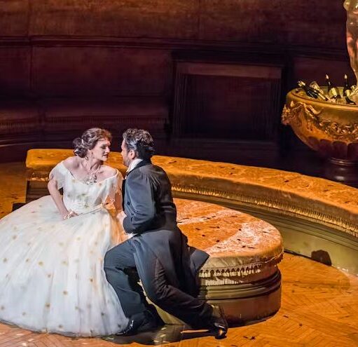 An opera scene showing a woman in a voluminous white gown sitting on a gold circular bench, looking intently at a man in a black suit kneeling before her, with a large, ornate golden bowl and statue in the background.