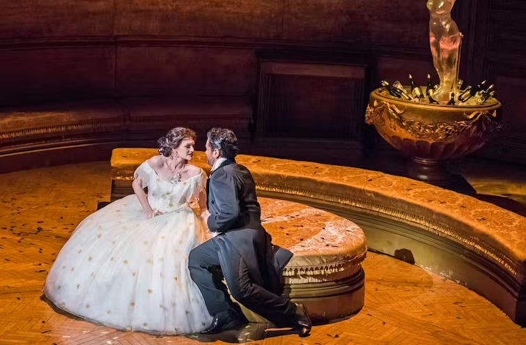 An opera scene showing a woman in a voluminous white gown sitting on a gold circular bench, looking intently at a man in a black suit kneeling before her, with a large, ornate golden bowl and statue in the background.