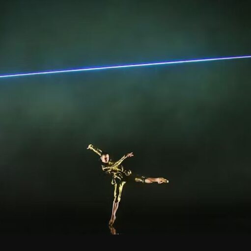 A dancer wearing a shimmering gold costume is frozen mid-pose, standing on the right leg with the left leg extended backward, illuminated by a horizontal blue-violet neon light strip crossing the top half of the dark stage.