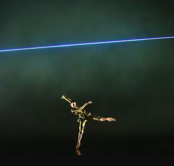 A dancer wearing a shimmering gold costume is frozen mid-pose, standing on the right leg with the left leg extended backward, illuminated by a horizontal blue-violet neon light strip crossing the top half of the dark stage.