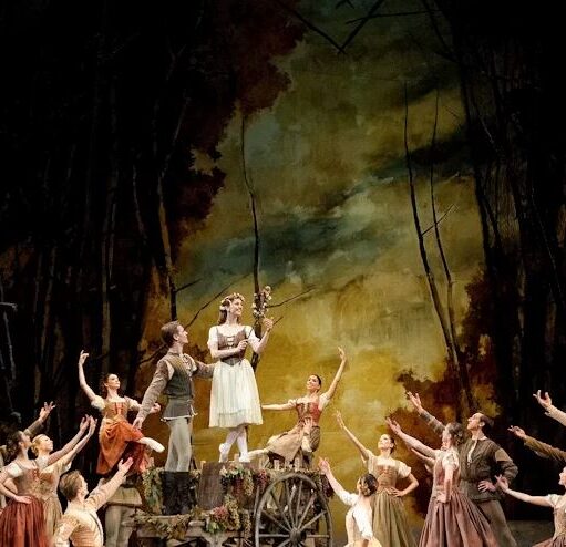 A large group of ballet dancers in rustic costumes surround a woman standing on a wooden cart holding a small bouquet of flowers, against a dark painted backdrop resembling a forest.
