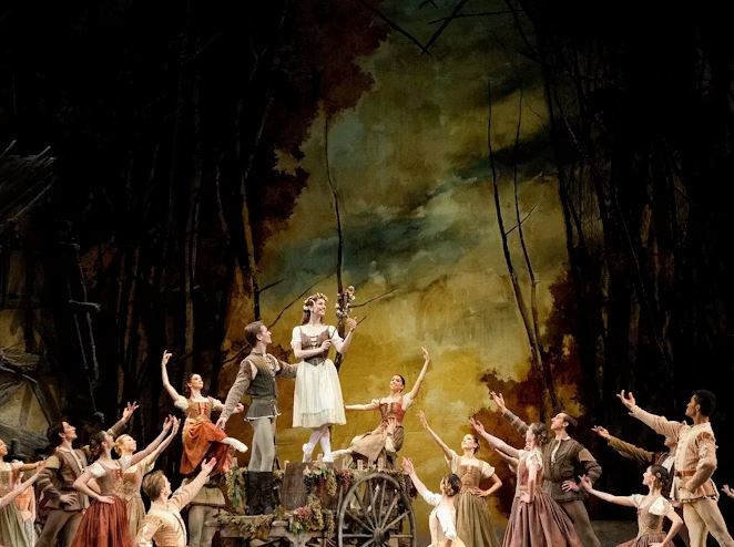 A large group of ballet dancers in rustic costumes surround a woman standing on a wooden cart holding a small bouquet of flowers, against a dark painted backdrop resembling a forest.