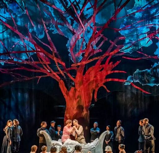 A theatre stage features a large, striking red tree with branching limbs against a dramatic backdrop of blue and black lighting. In the center, two figures in lighter clothing sit together on a white bed surrounded by a chorus of people dressed in dark clothing.