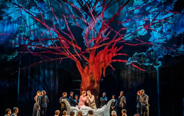A theatre stage features a large, striking red tree with branching limbs against a dramatic backdrop of blue and black lighting. In the center, two figures in lighter clothing sit together on a white bed surrounded by a chorus of people dressed in dark clothing.