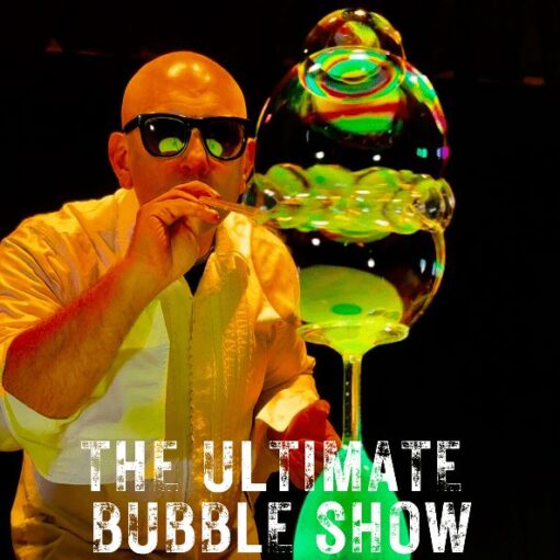 A brightly-lit photograph of a bald man in a yellow shirt and black sunglasses blowing bubbles with a wand, creating a large, colorful bubble structure that is glowing green. The text "THE ULTIMATE BUBBLE SHOW" is overlaid in white.