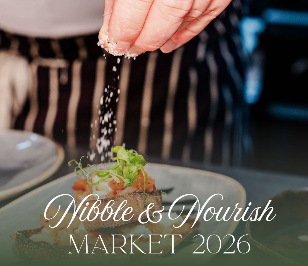 A close-up photograph of a chef's hand sprinkling white shavings, likely cheese, onto a small plated food item topped with garnish. White cursive text overlaid on the image reads "Nibble & Nourish" and "MARKET 2026" below it.