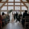 A photograph of a wedding ceremony taking place indoors. The bride and groom are standing before an officiant beneath a floral arch in front of a large, bright window. Guests are seated in white chairs on either side of the aisle.