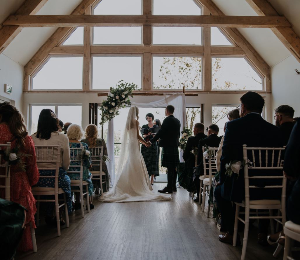A photograph of a wedding ceremony taking place indoors. The bride and groom are standing before an officiant beneath a floral arch in front of a large, bright window. Guests are seated in white chairs on either side of the aisle.