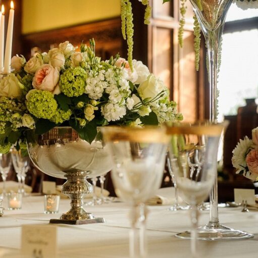 A close-up photograph of a lavishly set dining table with a silver bowl centerpiece full of white, light pink, and lime green flowers, surrounded by silver candlesticks and delicate glassware with gold rims.