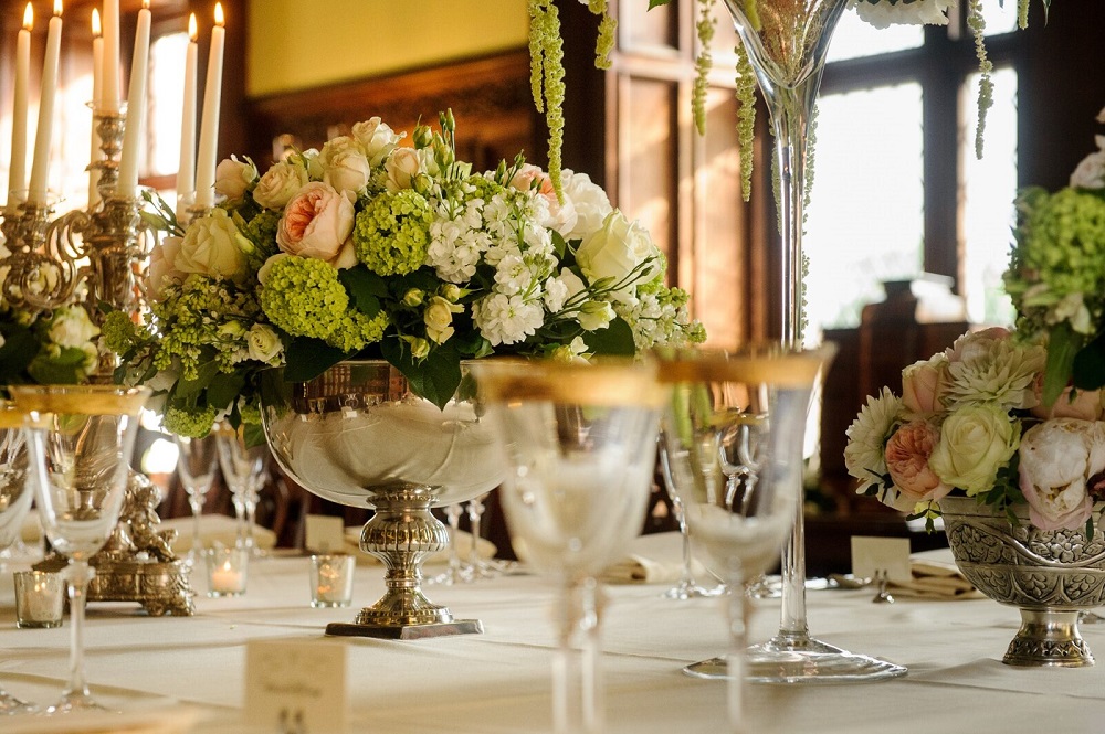 A close-up photograph of a lavishly set dining table with a silver bowl centerpiece full of white, light pink, and lime green flowers, surrounded by silver candlesticks and delicate glassware with gold rims.