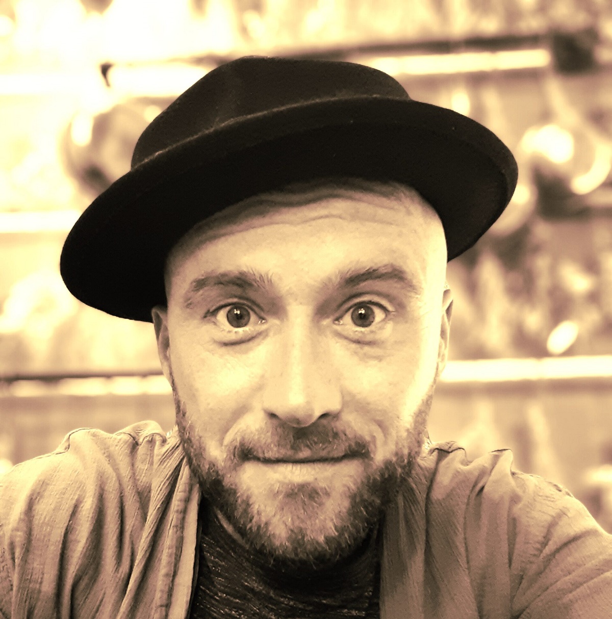 A sepia-toned, close-up photograph of a smiling, bearded man with a shaved head, wearing a dark fedora hat and a jacket.