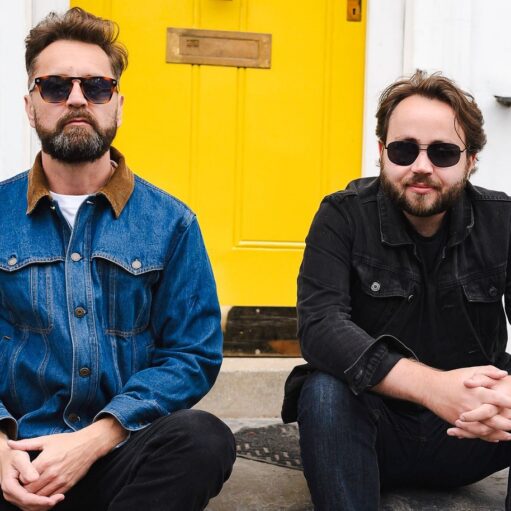 A photograph of two bearded men sitting on a doorstep in front of a bright yellow door. The man on the left is wearing a denim jacket and sunglasses, and the man on the right is wearing a black denim jacket and sunglasses.