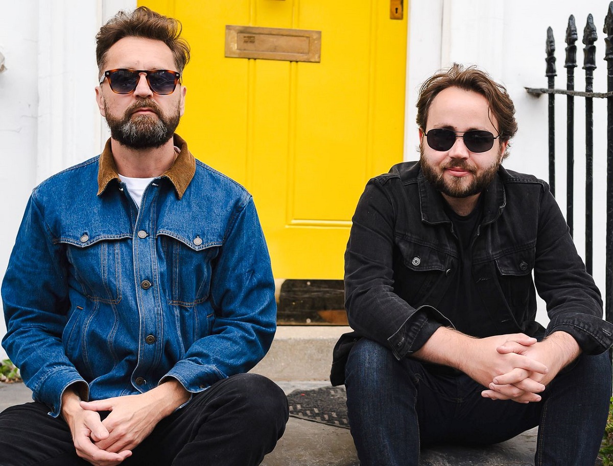 A photograph of two bearded men sitting on a doorstep in front of a bright yellow door. The man on the left is wearing a denim jacket and sunglasses, and the man on the right is wearing a black denim jacket and sunglasses.