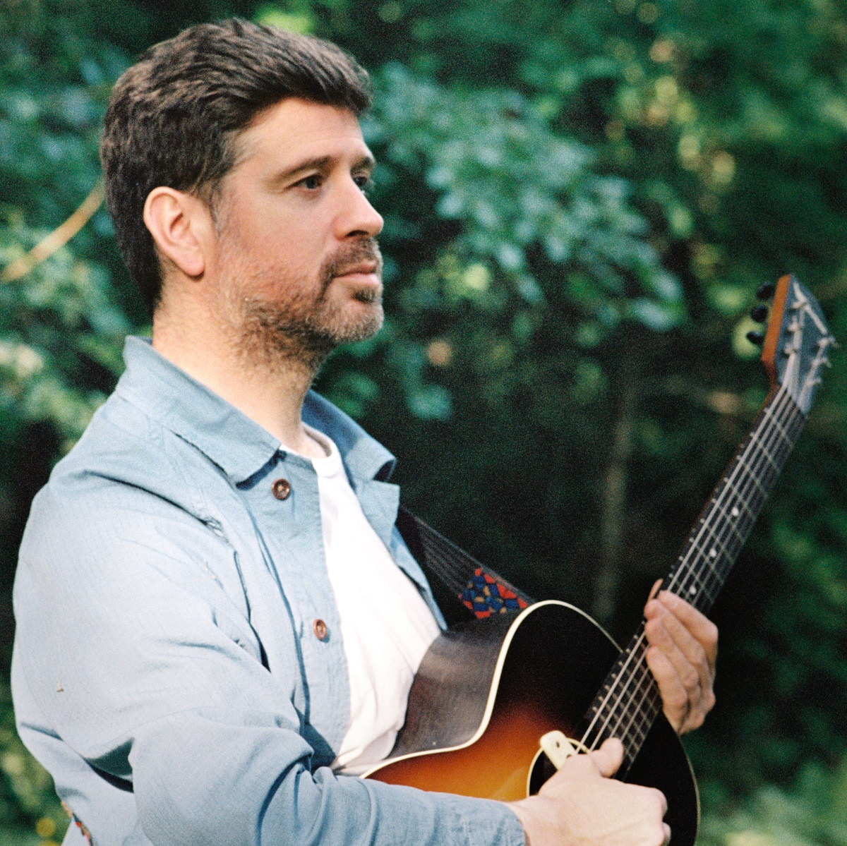 A portrait photograph of a man with dark hair and a beard, wearing a light blue collared shirt over a white shirt, holding a guitar. He is looking to the side against a backdrop of green foliage.