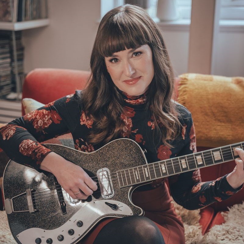 A photograph of a woman with long dark hair and bangs, sitting indoors with a black and silver electric guitar across her lap, looking at the camera. She is wearing a floral dark top and an orange-red skirt.
