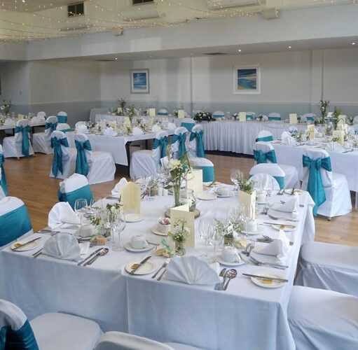 A photograph of a large, well-lit indoor wedding reception setup with square tables covered in white linens and white chairs. The chairs have teal/turquoise sashes and bows, and the tables are set with white napkins, simple centerpieces, and white floral arrangements.