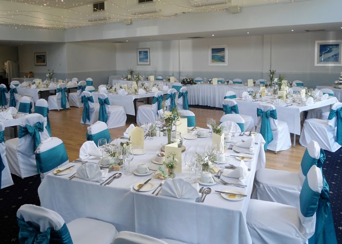 A photograph of a large, well-lit indoor wedding reception setup with square tables covered in white linens and white chairs. The chairs have teal/turquoise sashes and bows, and the tables are set with white napkins, simple centerpieces, and white floral arrangements.