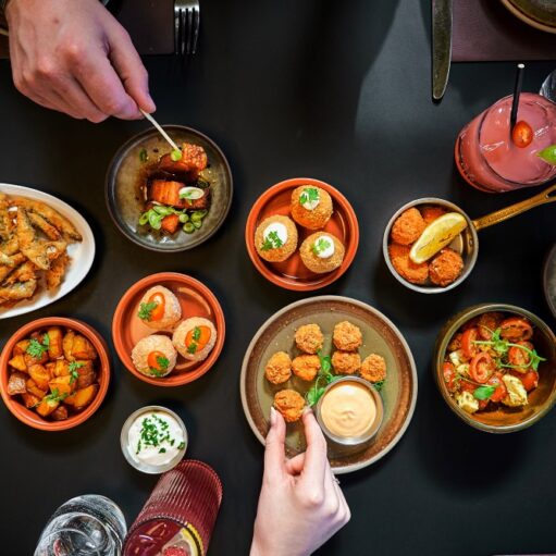 A high-angle photograph showing a black table filled with various small dishes of tapas being shared by people, including crispy fried fish, patatas bravas, croquettes, spring rolls, and a pink cocktail.
