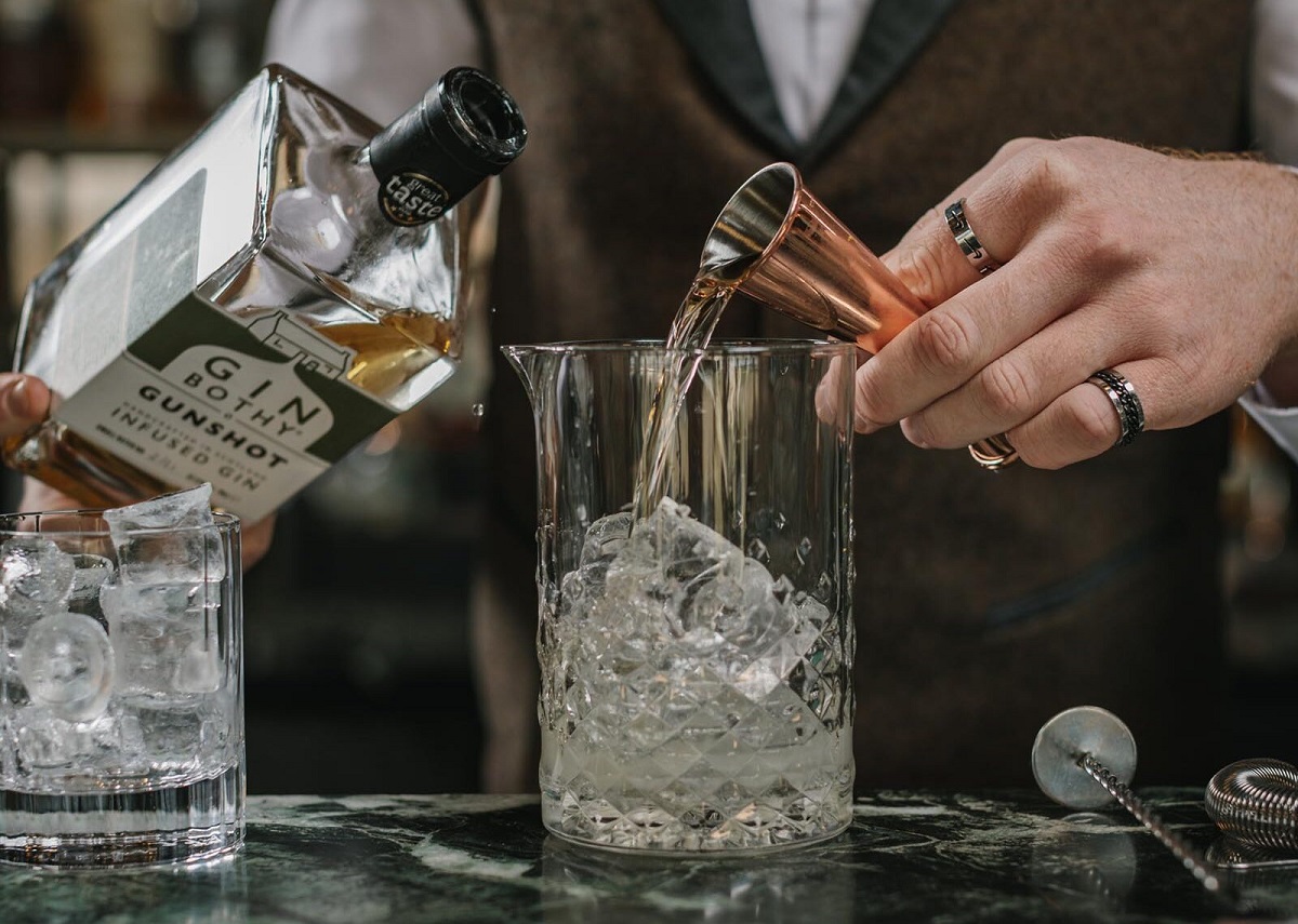 A close-up photograph of a bartender pouring Gin Bothy Gunshot Infused Gin from a copper jigger into a mixing glass filled with ice and liquid, while a bottle of the gin is held in the background.