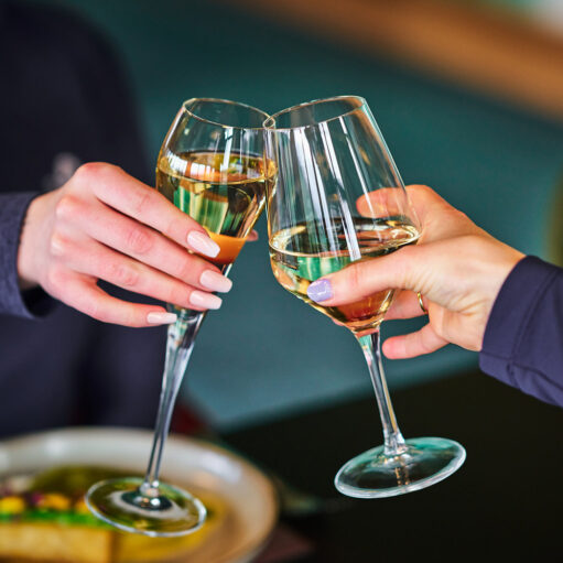 A close-up photograph of two people making a toast, clinking two fluted wine glasses filled with a light-colored beverage, possibly white wine or champagne, over a blurred plate of food.