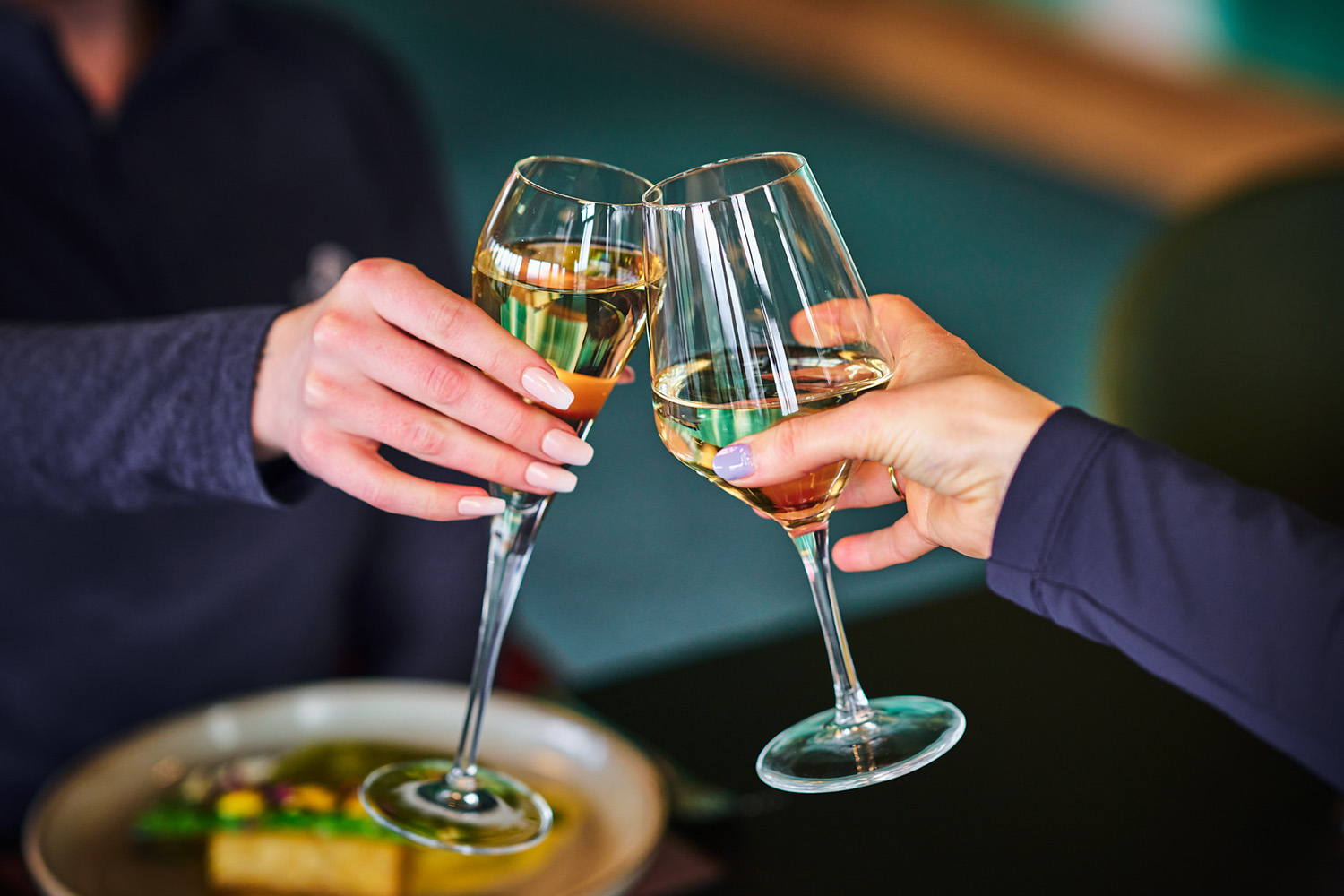 A close-up photograph of two people making a toast, clinking two fluted wine glasses filled with a light-colored beverage, possibly white wine or champagne, over a blurred plate of food.