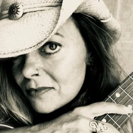 A close-up, black and white photo of a woman wearing a straw cowboy hat that partially shades her face. She has a nose ring, is looking directly at the camera, and is holding the fretboard of a stringed instrument (likely a guitar or ukulele) against her shoulder. She is also wearing a prominent ring on her finger.