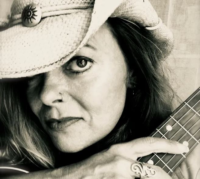 A close-up, black and white photo of a woman wearing a straw cowboy hat that partially shades her face. She has a nose ring, is looking directly at the camera, and is holding the fretboard of a stringed instrument (likely a guitar or ukulele) against her shoulder. She is also wearing a prominent ring on her finger.