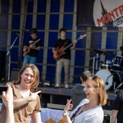 Two women smiling and pointing towards a music stage where two male musicians are playing electric guitars/basses. The stage backdrop is blue and features a logo that says "MONTROSE MUSIC". The photo appears to be taken at an outdoor music festival or event.