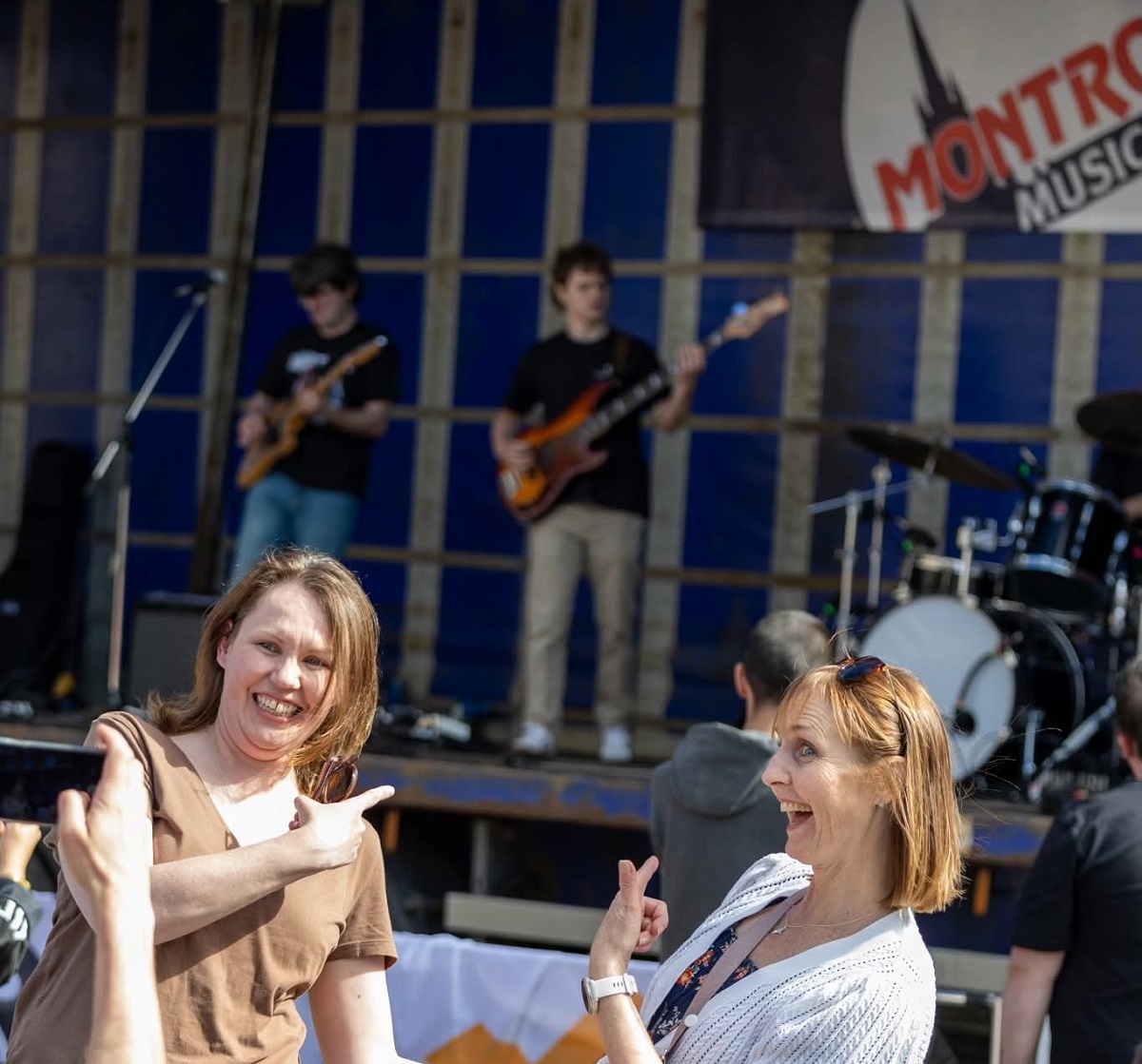 Two women smiling and pointing towards a music stage where two male musicians are playing electric guitars/basses. The stage backdrop is blue and features a logo that says "MONTROSE MUSIC". The photo appears to be taken at an outdoor music festival or event.