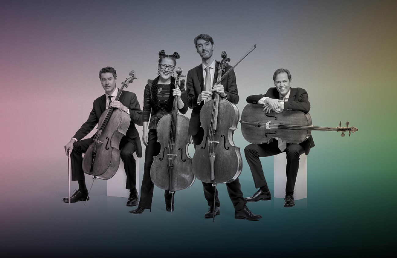 A black-and-white photograph of a four-person cello ensemble posed against a vibrant, multi-colored gradient background. Two musicians are seated and two are standing, all holding their cellos, dressed in formal dark attire.