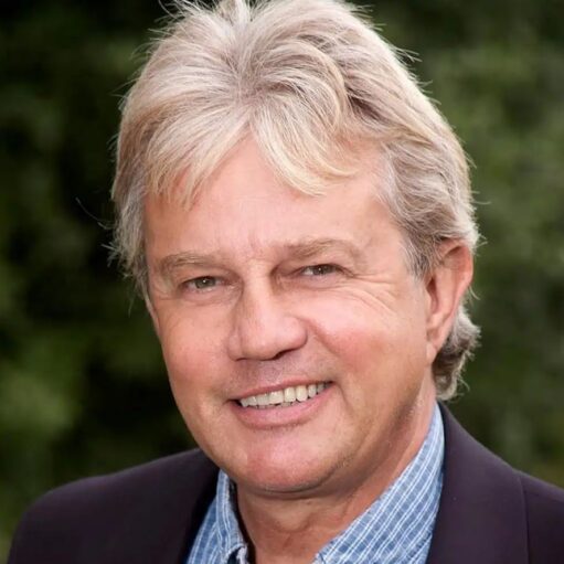 A headshot of a smiling man with graying blonde hair and blue eyes, wearing a dark jacket over a blue and white checkered shirt. The background is a soft, out-of-focus green.