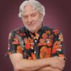 A close-up portrait of a smiling man with white hair and a white beard, wearing a dark blue floral-print shirt. The background is a soft-focus greenery, creating a bright and natural feel.