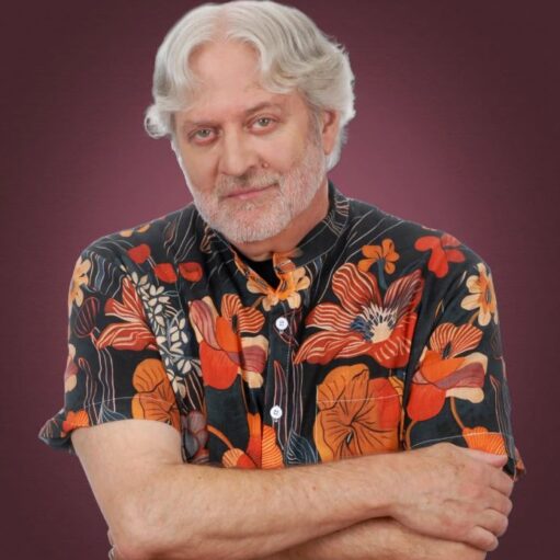 A close-up portrait of a smiling man with white hair and a white beard, wearing a dark blue floral-print shirt. The background is a soft-focus greenery, creating a bright and natural feel.
