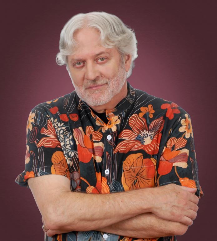 A close-up portrait of a smiling man with white hair and a white beard, wearing a dark blue floral-print shirt. The background is a soft-focus greenery, creating a bright and natural feel.