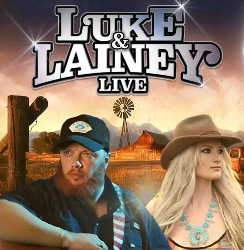 A promotional graphic for "Luke & Lainey Live" featuring two performers in front of a rural landscape. On the left is a man with a beard wearing a black baseball cap and a dark shirt. On the right is a woman with long blonde hair wearing a wide-brimmed brown hat and a turquoise necklace. The background shows a wooden fence, a windmill, a barn, and distant mountains under a warm sunset sky.