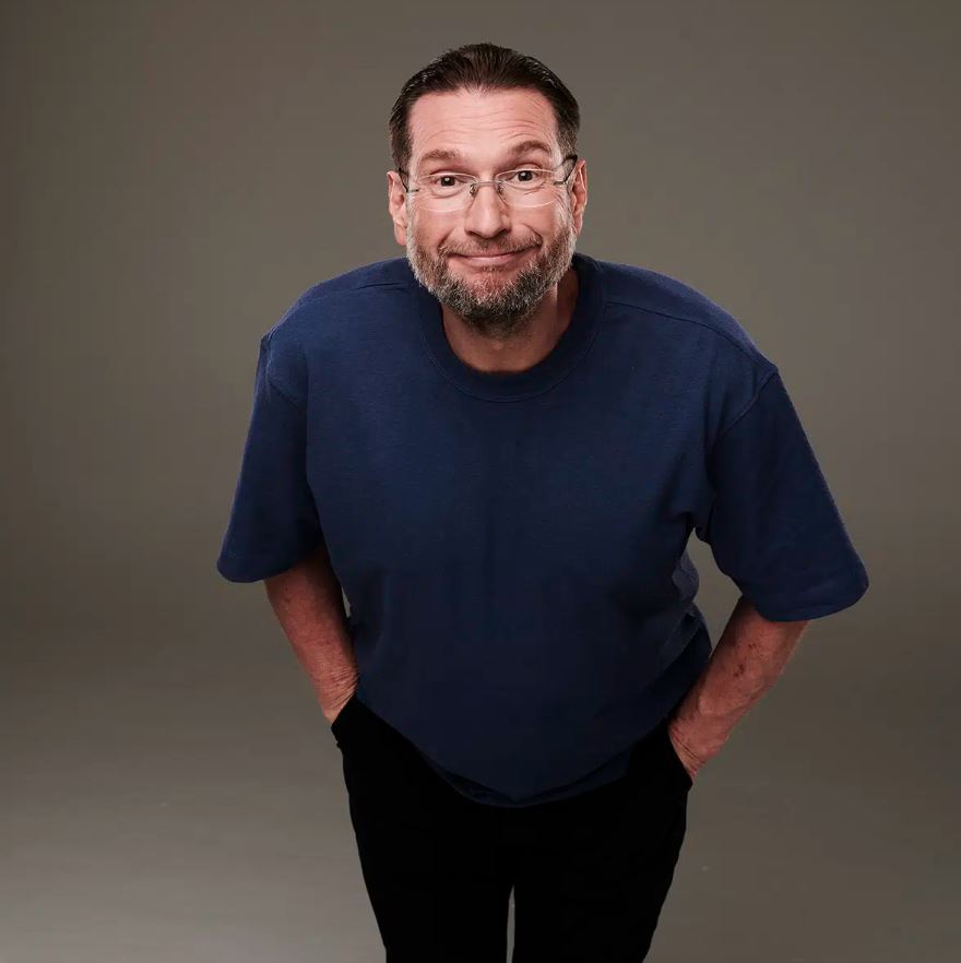 A high-angle studio portrait of a man with short brown hair, a groomed beard, and glasses, wearing a navy blue t-shirt and black trousers. He is leaning slightly toward the camera with his hands in his pockets, sporting a playful, wide-eyed smirk against a neutral gray background.