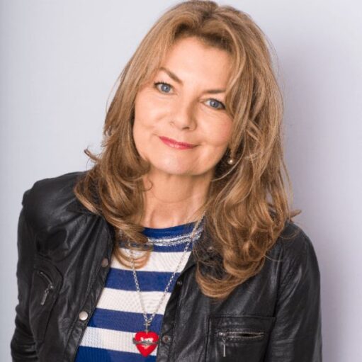 A close-up studio headshot of a woman with long, layered strawberry-blonde hair and blue eyes, smiling softly at the camera. She is wearing a black leather jacket over a blue and white striped top and a silver necklace with a prominent red heart pendant. The photo is set against a clean, light gray background.