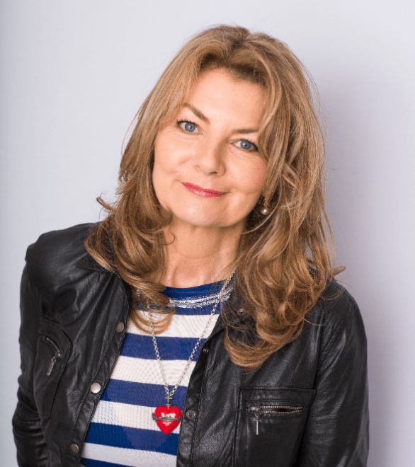 A close-up studio headshot of a woman with long, layered strawberry-blonde hair and blue eyes, smiling softly at the camera. She is wearing a black leather jacket over a blue and white striped top and a silver necklace with a prominent red heart pendant. The photo is set against a clean, light gray background.