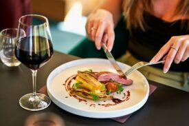 A close-up, shallow-focus shot of a person at a dinner table cutting into a seared duck breast served with roasted endive and a balsamic reduction. A glass of red wine stands in the foreground on the left.