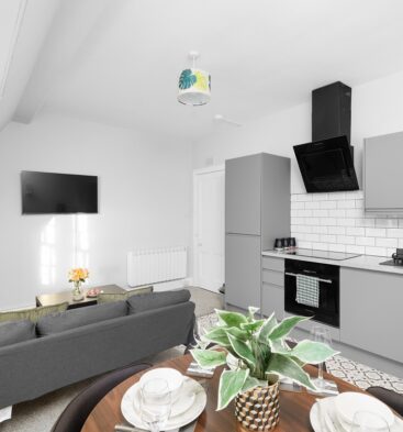 An open-plan living area and kitchen featuring sleek grey cabinets, a white subway tile backsplash, and a black oven and extractor fan. A grey sofa faces a wall-mounted TV, and a dining table with place settings is visible in the foreground.