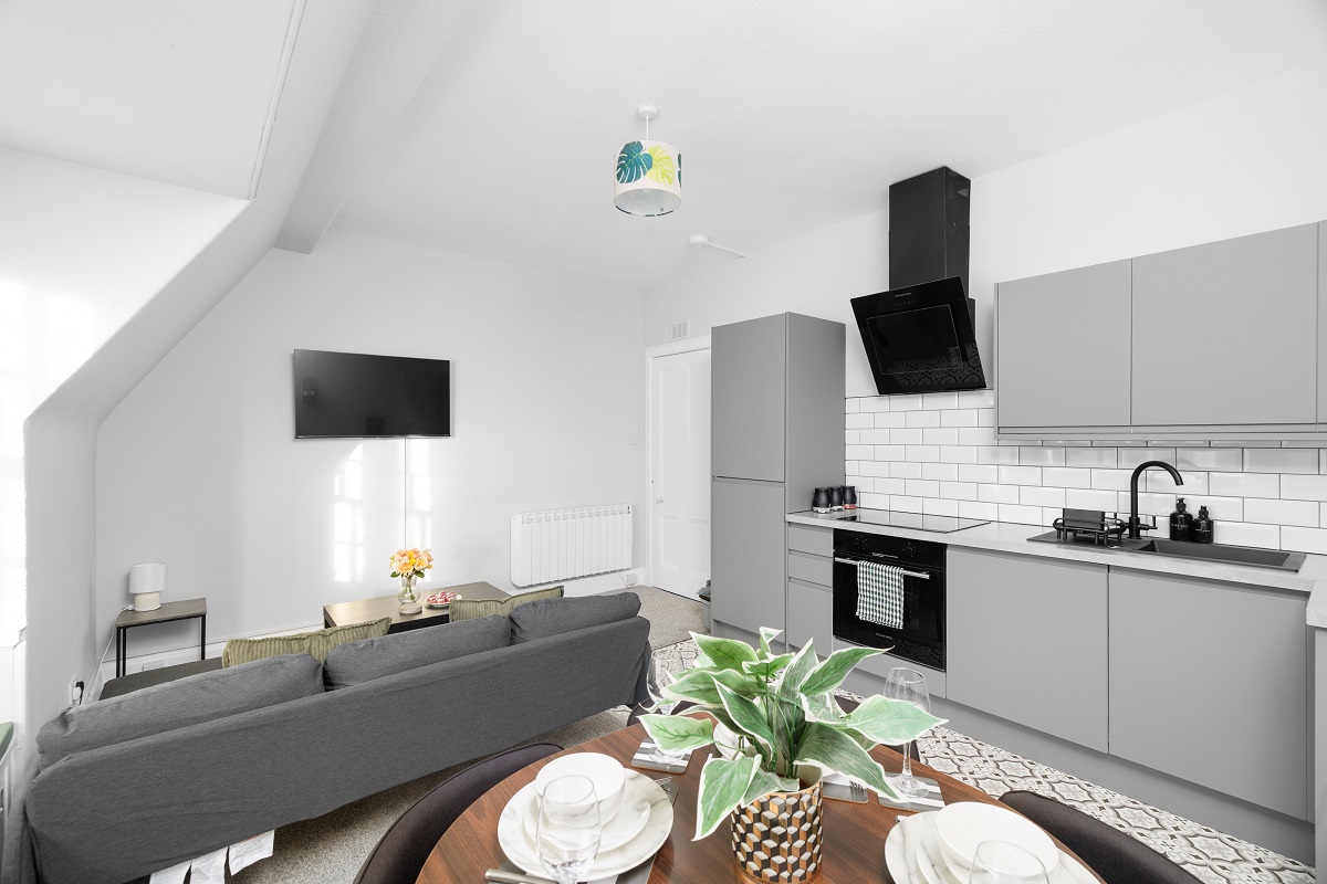An open-plan living area and kitchen featuring sleek grey cabinets, a white subway tile backsplash, and a black oven and extractor fan. A grey sofa faces a wall-mounted TV, and a dining table with place settings is visible in the foreground.