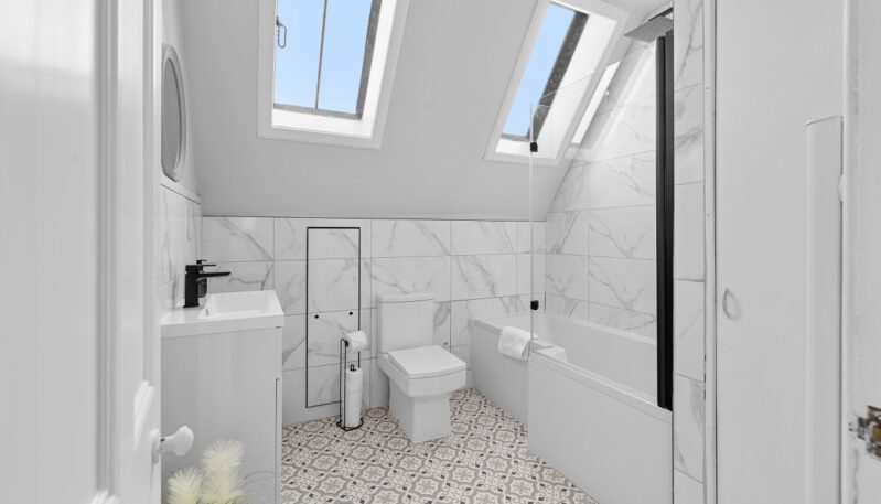 A bright, modern bathroom featuring white marble-style wall tiles, patterned floor tiles, and a white vanity with a black faucet. The room has a sloped ceiling with two skylights illuminating a combination bathtub and shower with a glass screen.